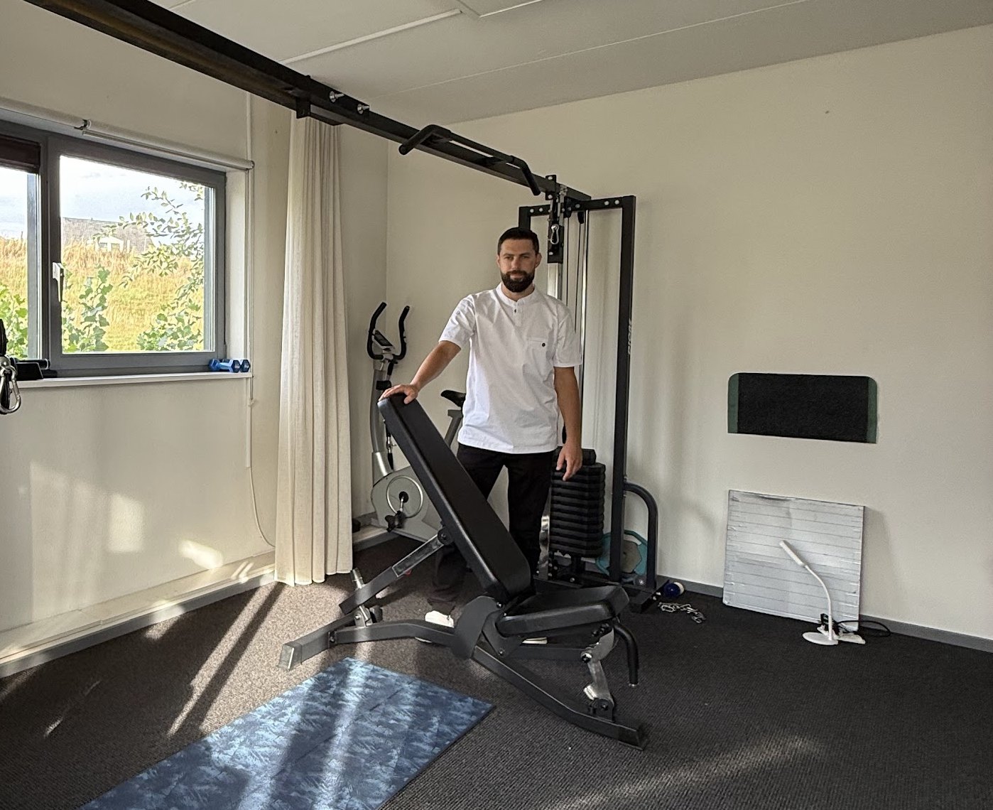 De oefenruimte met de Bubnovsky-trainer, een trainingsbank en een blauwe mat; Maxim staat bij de trainer